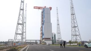 an orange crane sits above a large white building, surrounded by four towers of metal scaffolding