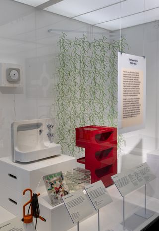 A vitrine at the V&A's new design galleries showing various household objects including the white compact porcelain sink from the Barbican apartments and Konstantin Grcic Mayday lamp for Flos, featuring an orange top with a black cable