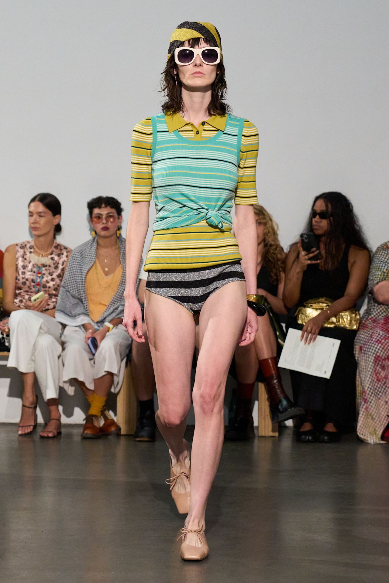 Layering staples for spring 2026 is shown in a photo from Zankov's runway show, with a model walking while wearing a bright green striped polo shirt layered underneath a teal striped polo shirt styled with a striped bikini brief that's styled with tan ballet flats, a black bangle, a printed scarf, and oversized white sunglasses