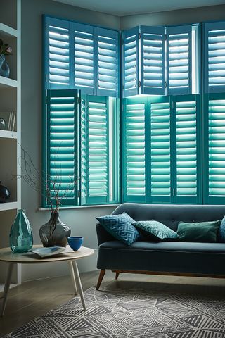 aquamarine blue shutters in living room with low couch, patterned cushions, rug and wooden coffee table topped with decorative glass vases