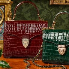 A deep red and deep green bag with gold detailing next to each other from Aspinal of London. 