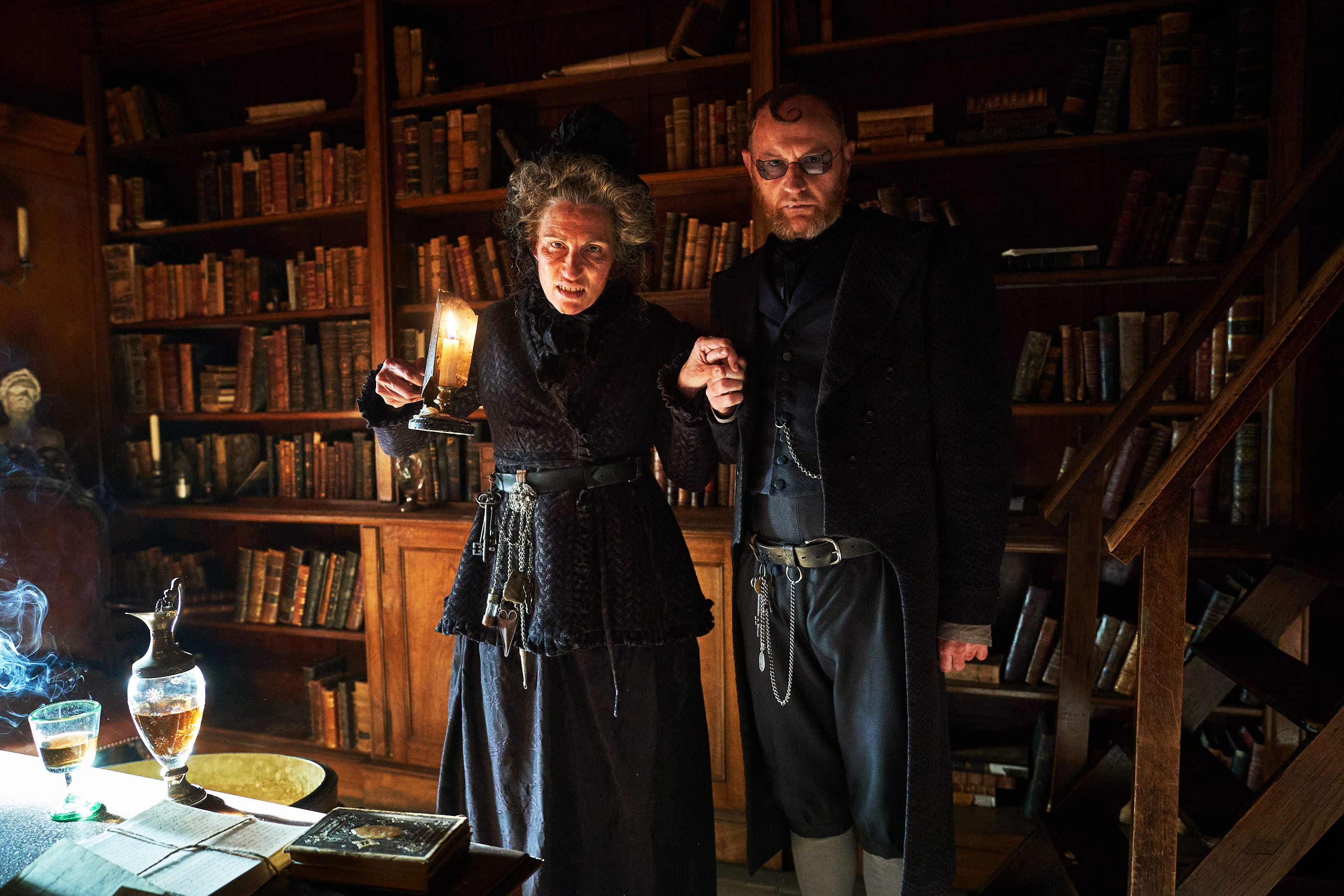 Mrs Wickens (Tamsin Greig) and Mr Wickens (Mark Gatiss) stand holding hands in the house's library, shelves behind them piled with books, while Mrs Wickens holds up a candle