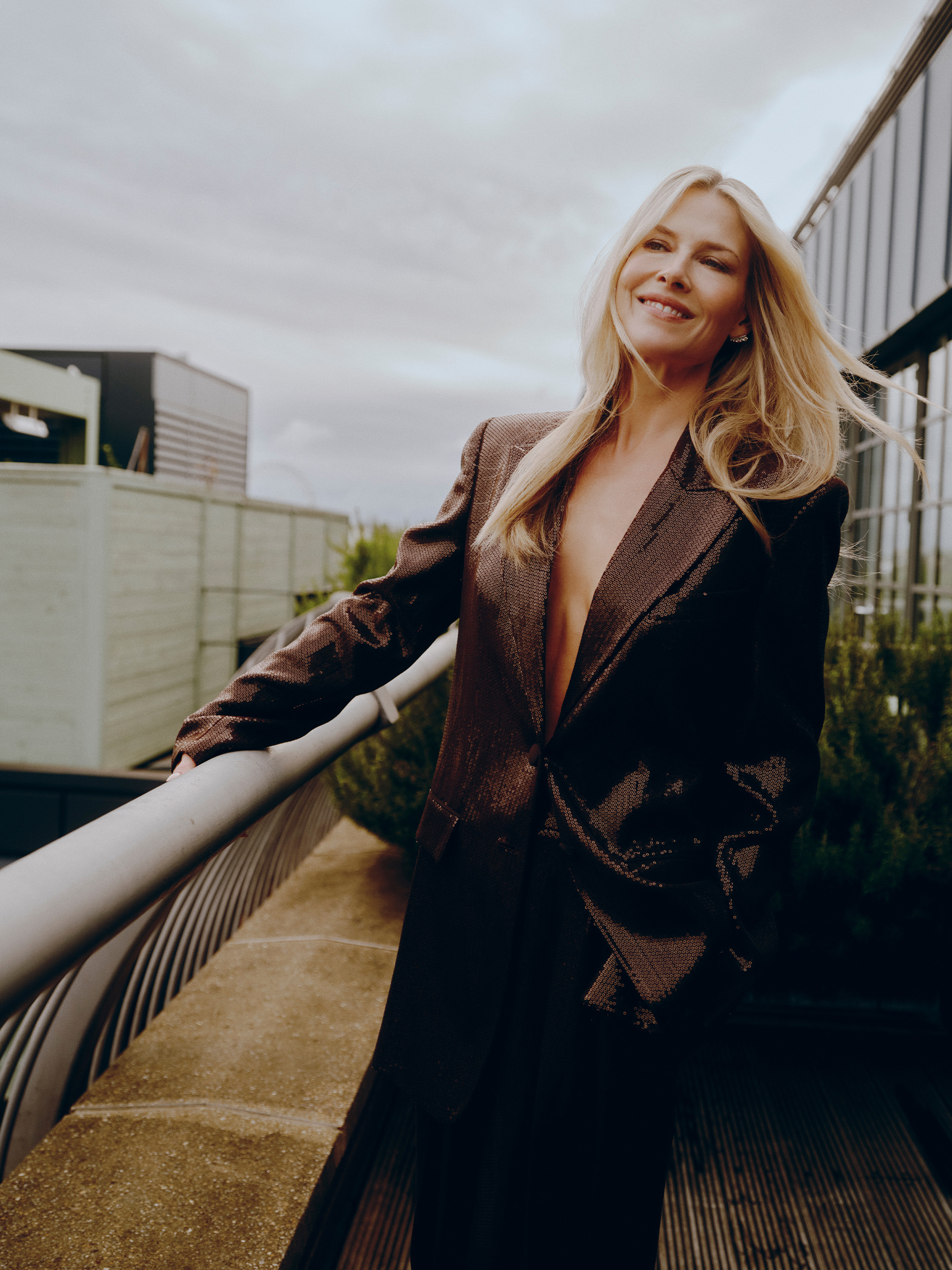 Ali Larter poses on the balcony of her London hotel. She is smiling with her hair blowing in the wind. She is wearing a chocolate brown sequin Michael Kors suit.