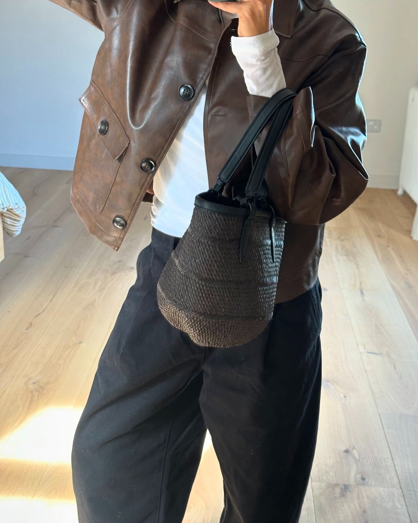 Marianne Smyth wears the best autumn jacket, a leather jacket. She pairs it with a long-sleeved whit top, black trousers a black raffia bag. A mirror picture, you can see her wooden floor and white walls in the back.