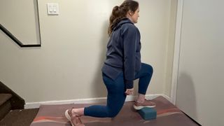 woman performing a forward lunge with one foot on a yoga block against a corridor wall