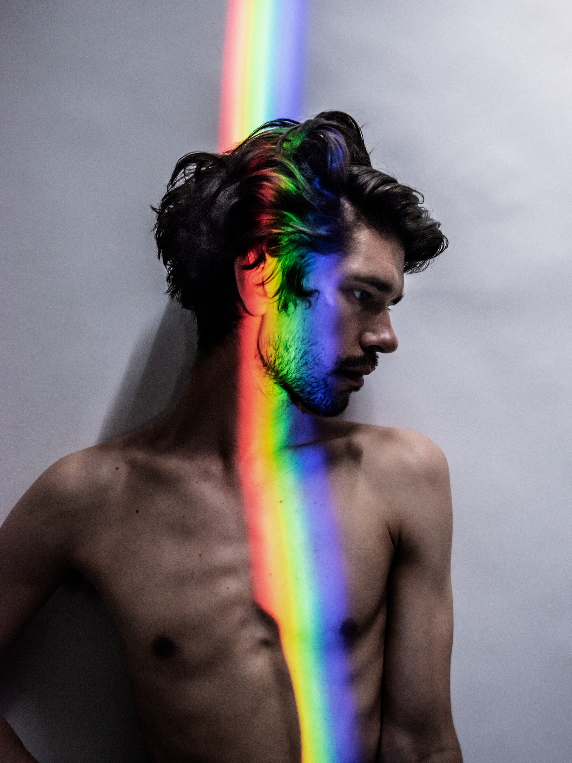 Colour photo of a shirtless Ben Whishaw leaning against a pale wall and looking to one side, with a vertical stripe of rainbow light from a prism falling across his face and chest.