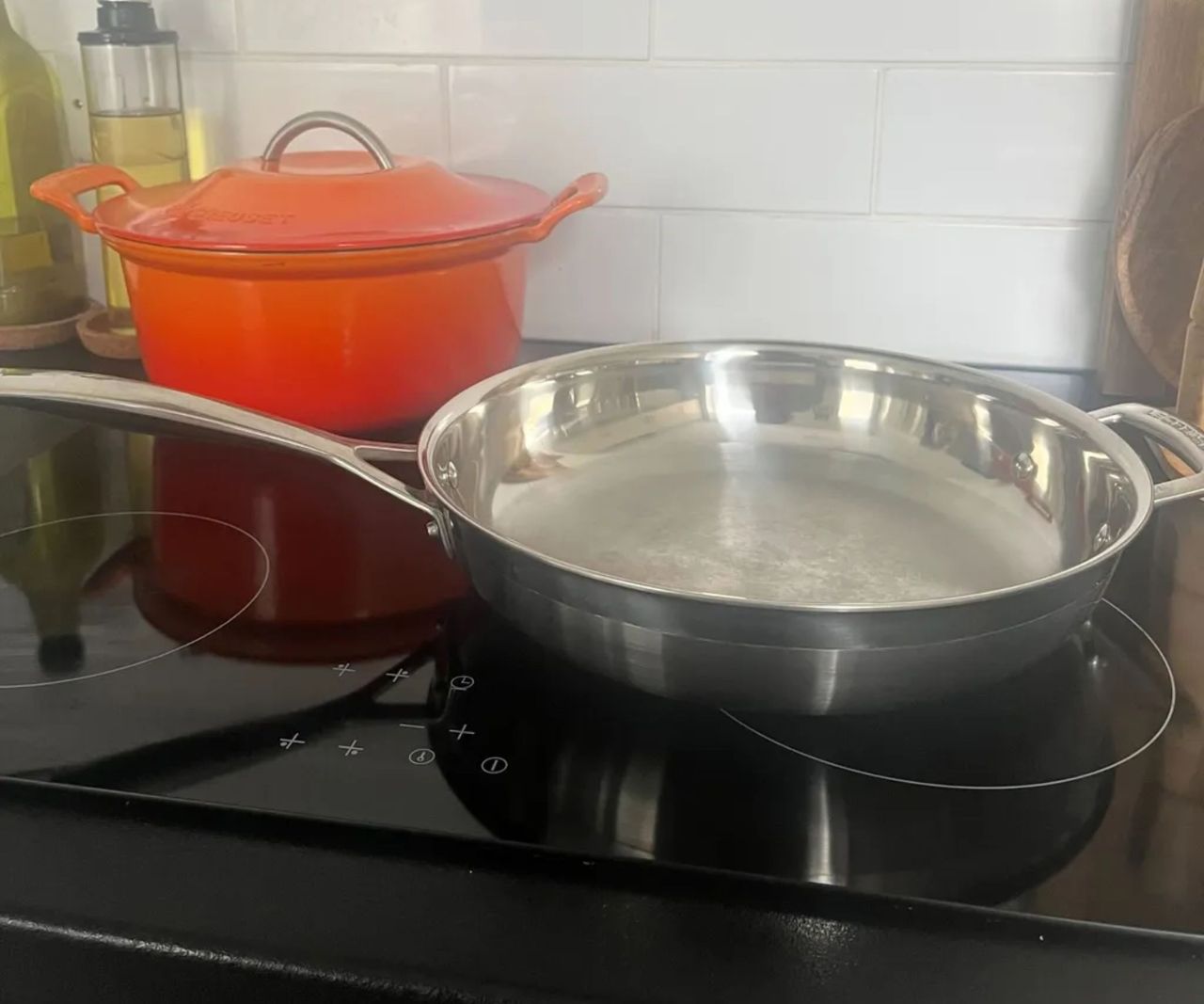 A stainless steel frying pan on a black stove in front of an orange Dutch oven.