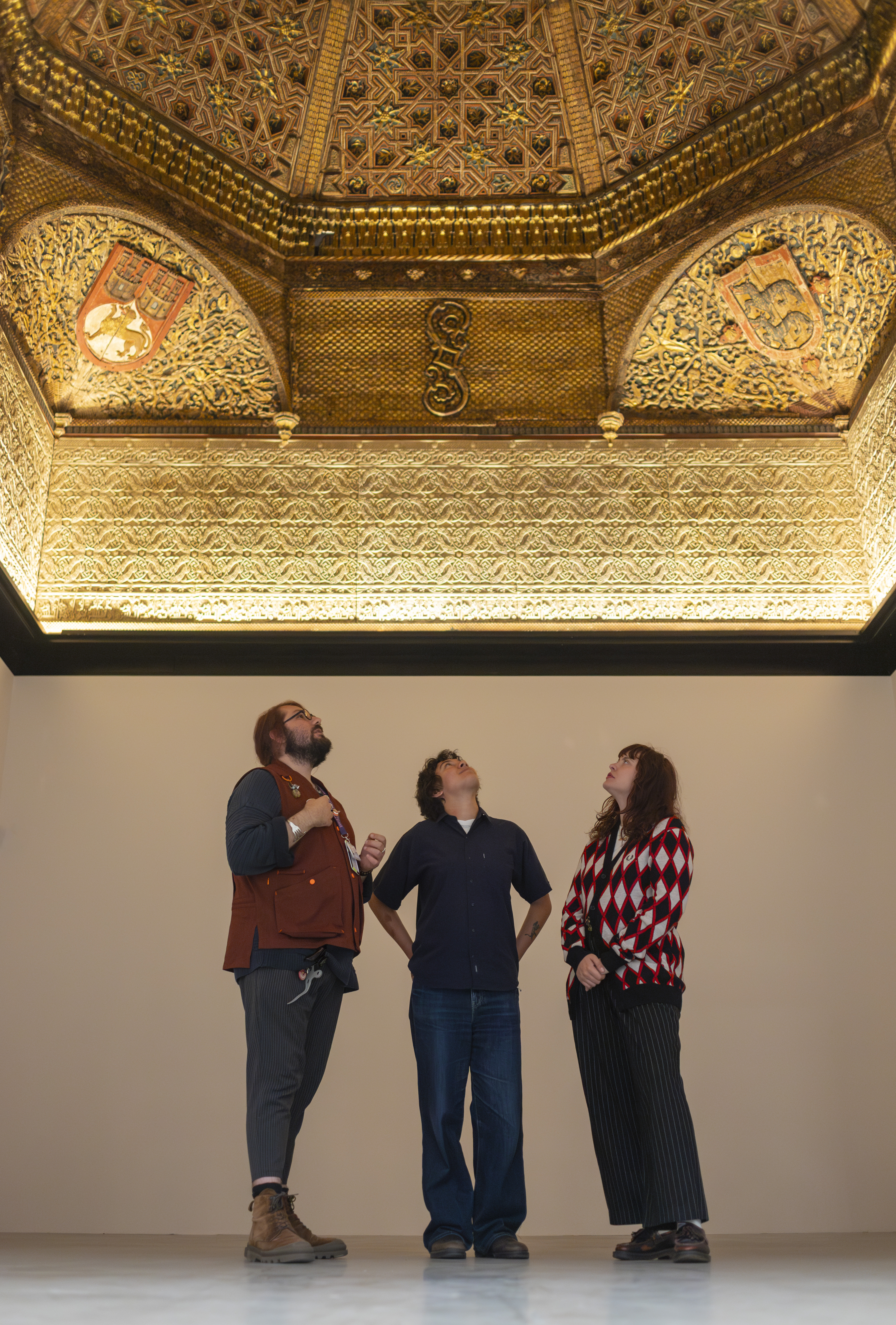 Three people gazing up at an ornate gilded 15th century ceiling from a Spanish palace, installed at the V&A East Storehouse