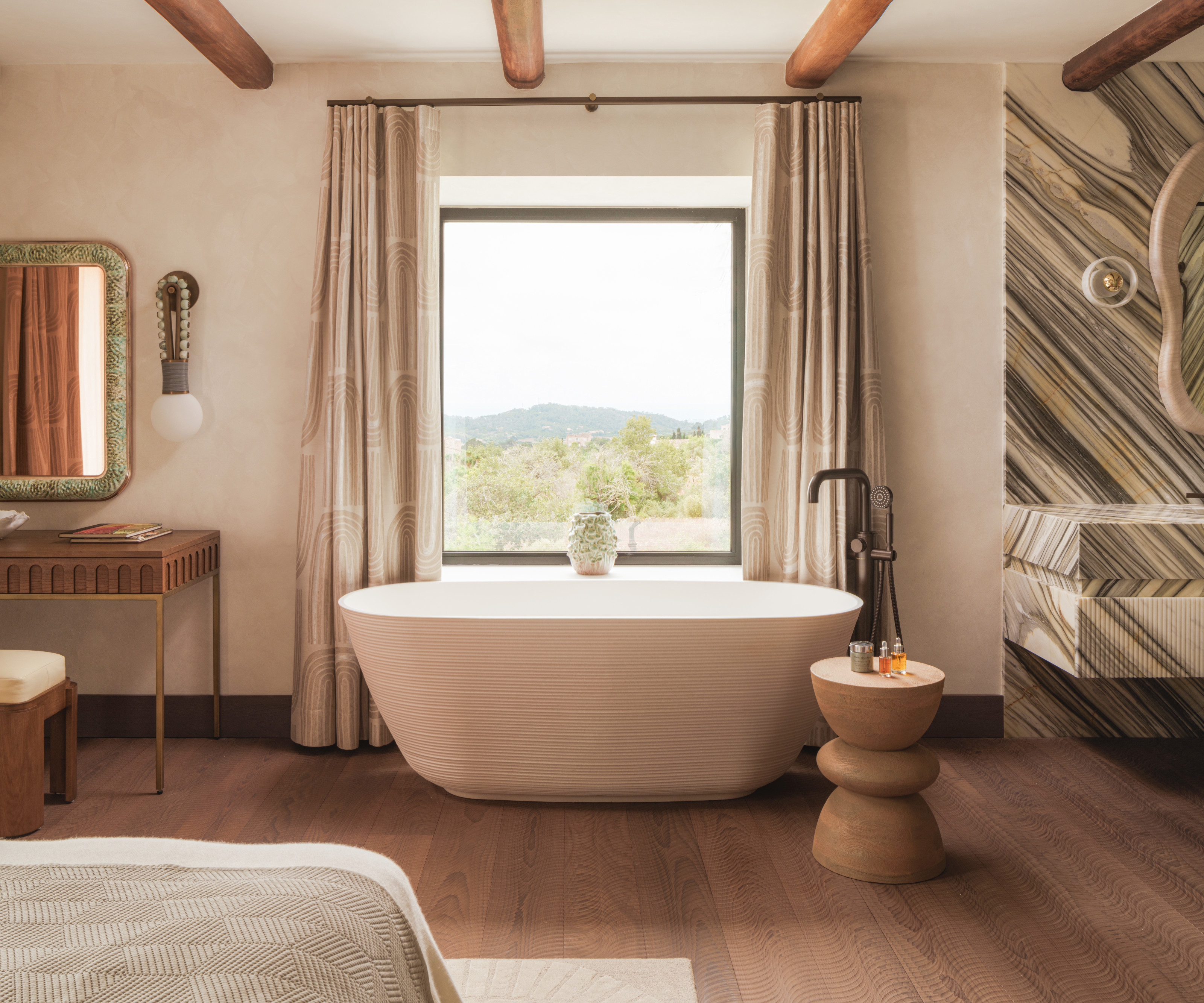 Textured white freestanding bathtub against picture window in bedroom with black floorstanding mixer, wood stool, and corner of vanity unit with grey and white striped marble