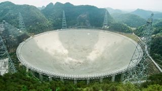 A large man-made dish sits like a crater surrounded by utility towers in a mountainous forest.