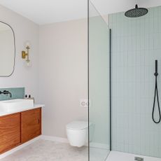 A modern bathroom with matt black showerhead and fitxture, teal green wall tiles, marble hexagonal floor tiles, teak wood vanity, rectangular ceramic bathroom basin, soft rectangular mirror, and pair of brass light fixtures on white walls