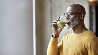 person wearing a yellow jumper drinking a glass of green smoothie while looking out through a window