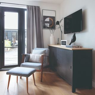 A living room with a TV hanging on the wall and a black sideboard placed below it with a chair and a matching footstool