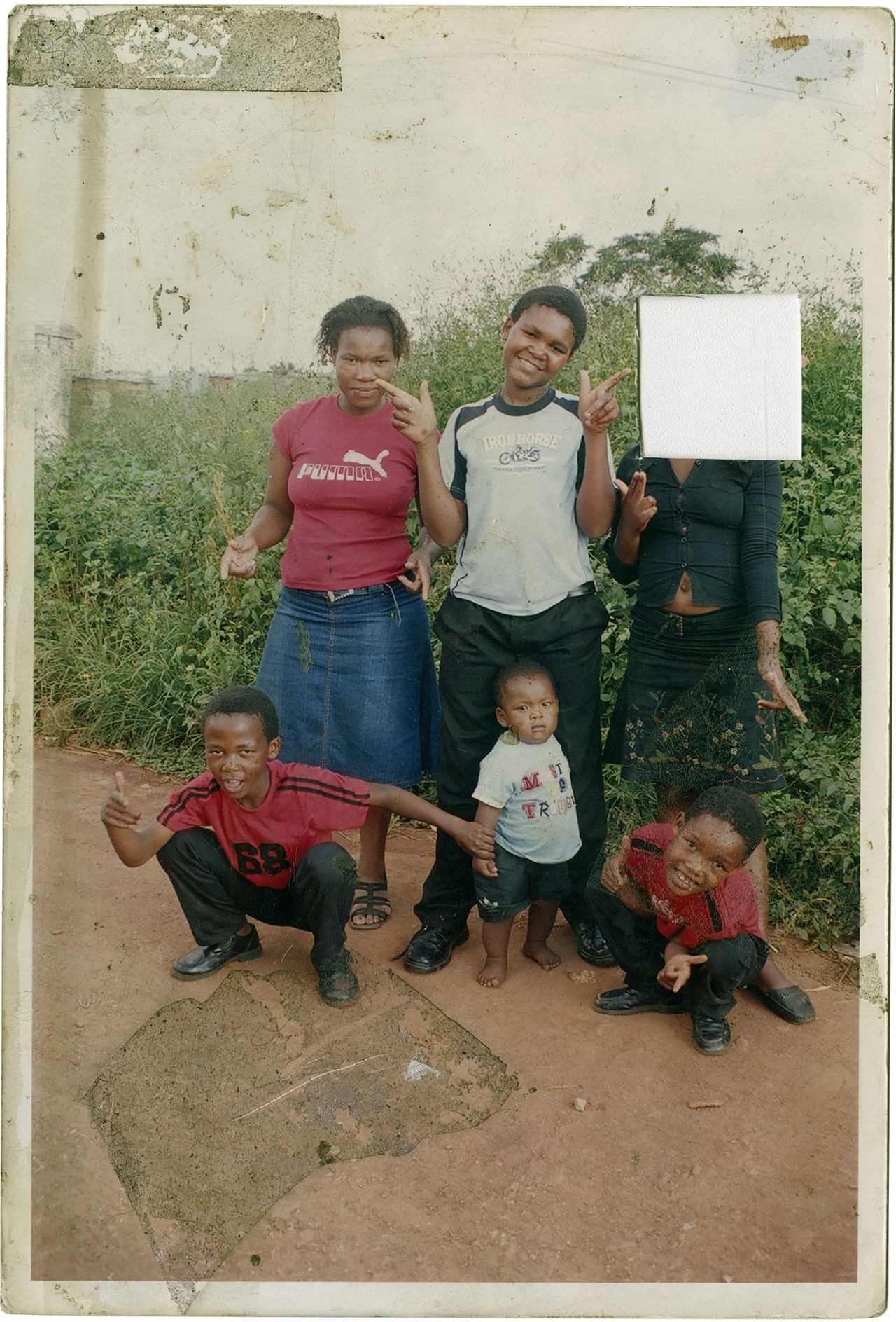 Color photograph of a five individuals. Two of the children are bent down while the toddler is stood between them. There are two older girls and a boy stood smiling for the camera. One of the girls&rsquo; face is obscured by a white square