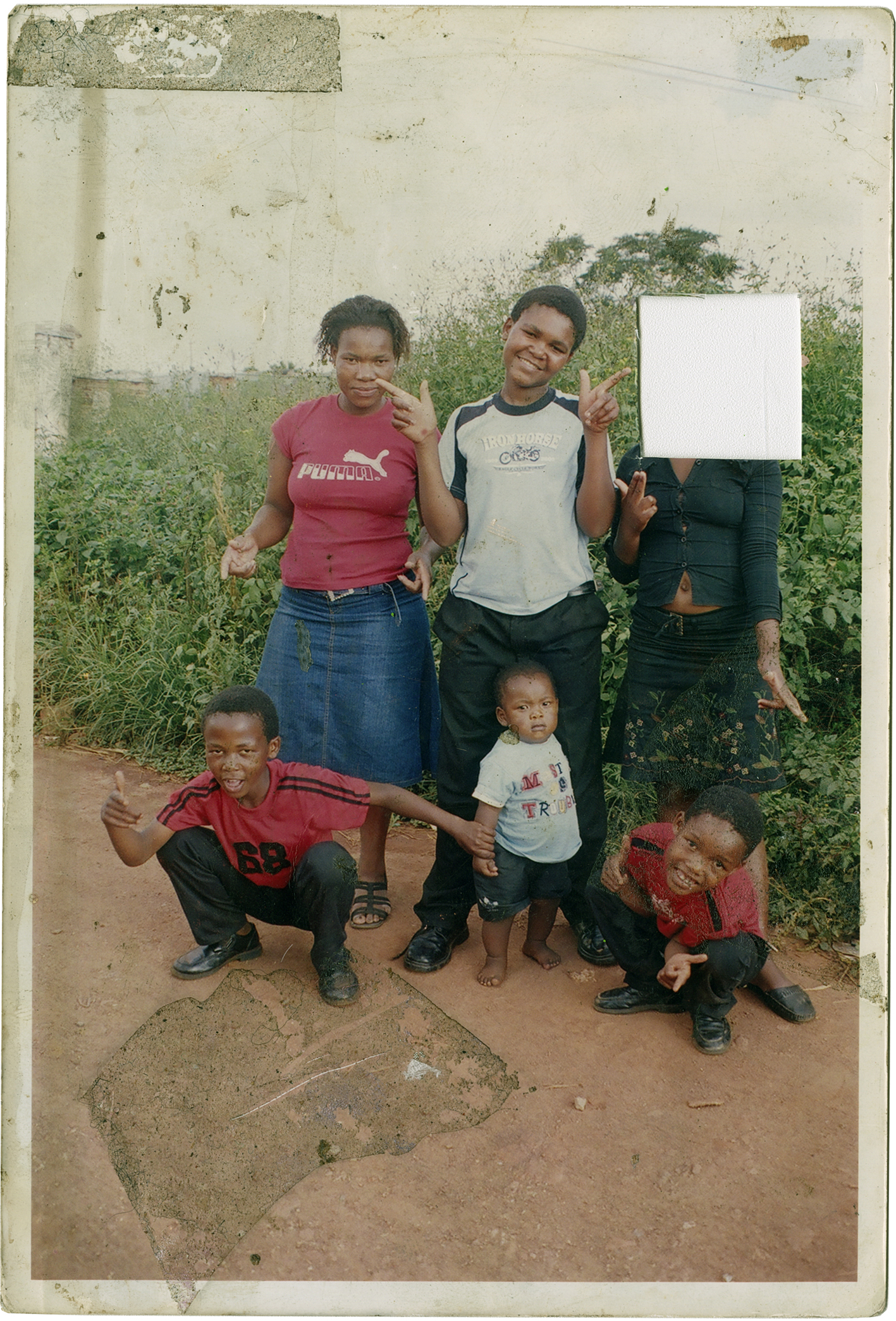 Color photograph of a five individuals. Two of the children are bent down while the toddler is stood between them. There are two older girls and a boy stood smiling for the camera. One of the girls’ face is obscured by a white square