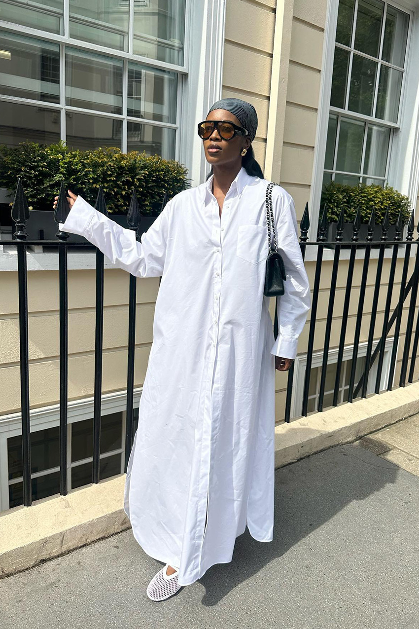 white outfit idea for summer is shown in a photo of a woman standing on the sidewalk in London wearing a navy blue headscarf with gold earrings, two-tone aviator sunglasses, a white maxi shirt dress, a black Chanel flap bag, and white mesh ballet flats
