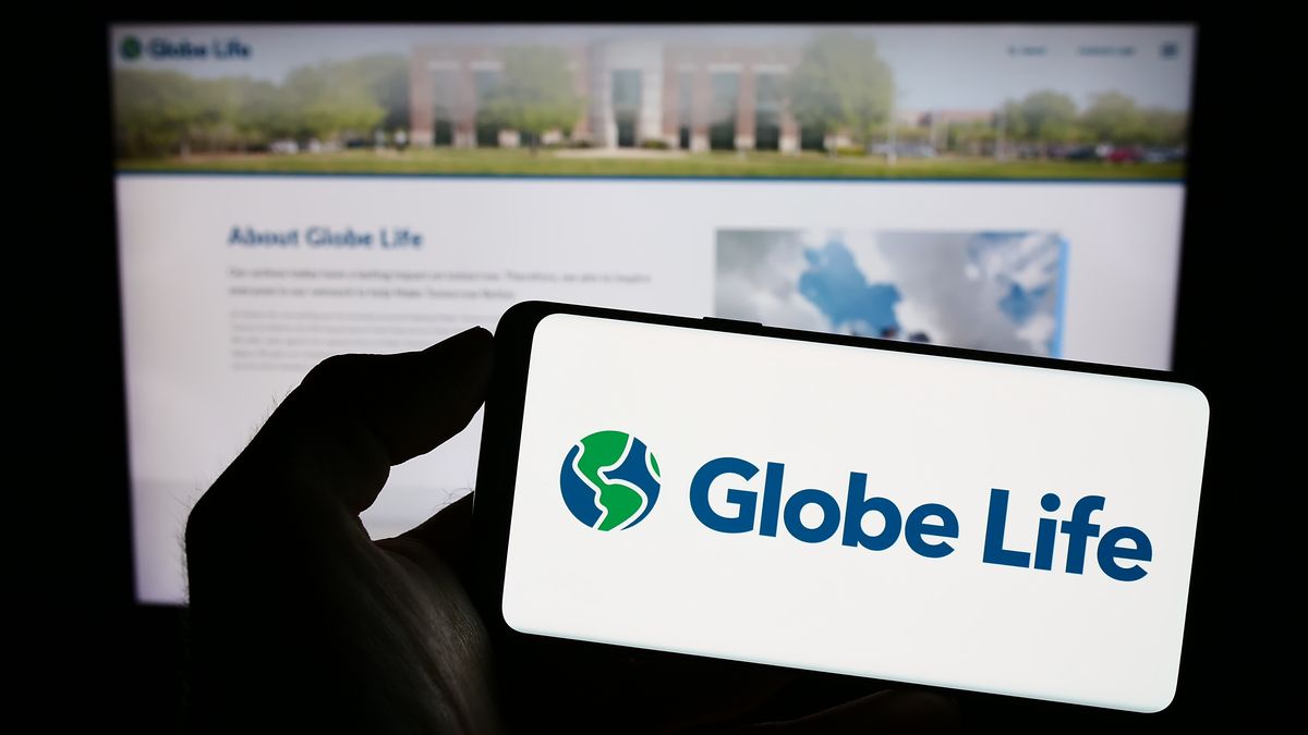 Globe Life insurance company logo on a cell phone in front of a monitor display the About page for the company. Shadowy hand holds the phone.