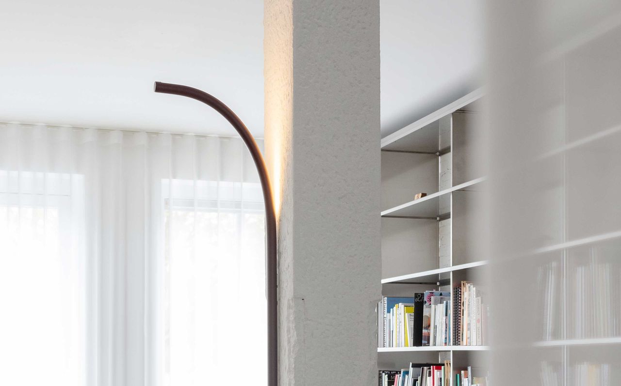 A living room with a bookcase in the background and a backlit rod-shaped wall lamp on a pillar