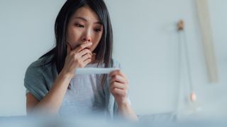 Asian woman looking at a pregnancy test, she has her hand over her mouth and looks thoughtful