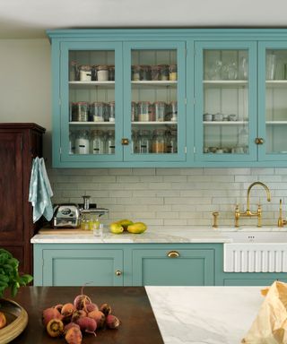 blue kitchen with freestanding antique storage unit, glazed upper cabinetry, pearlescent backsplash tiles, marble countertop, fluted sink, brass taps, island to fore, deVOL