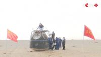A team of people examine a metal spacecraft in the middle of a sandy area with Chinese flags waving nearby
