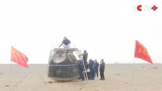 A team of people examine a metal spacecraft in the middle of a sandy area with Chinese flags waving nearby