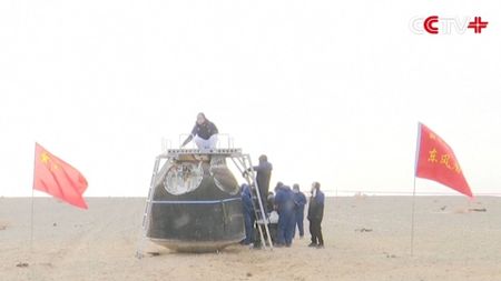 A team of people examine a metal spacecraft in the middle of a sandy area with Chinese flags waving nearby