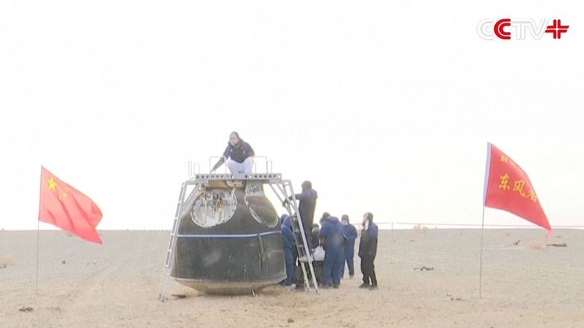 A team of people examine a metal spacecraft in the middle of a sandy area with Chinese flags waving nearby