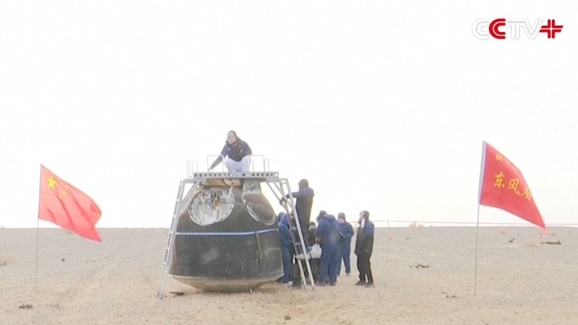 A team of people examine a metal spacecraft in the middle of a sandy area with Chinese flags waving nearby
