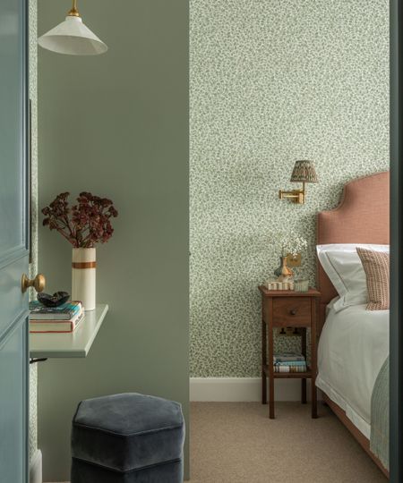 pale sage green bedroom with floral wallpaper, a pink upholstered bed, little wooden antique nightstand and a small desk shelf with a navy velvet stool