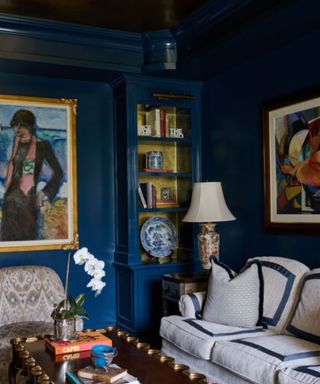 A dark blue living room with a blue and white couch, bookshelves and artwork