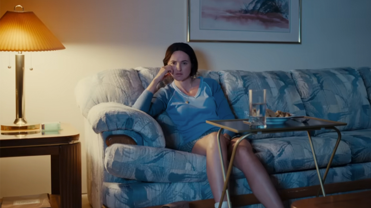 Renate Reinsve sitting on a blue couch with a TV dinner stand in front of her in Backrooms.
