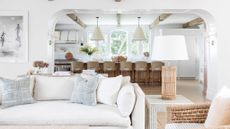 living room in white with white sofa and kitchen in background with island and rattan chairs