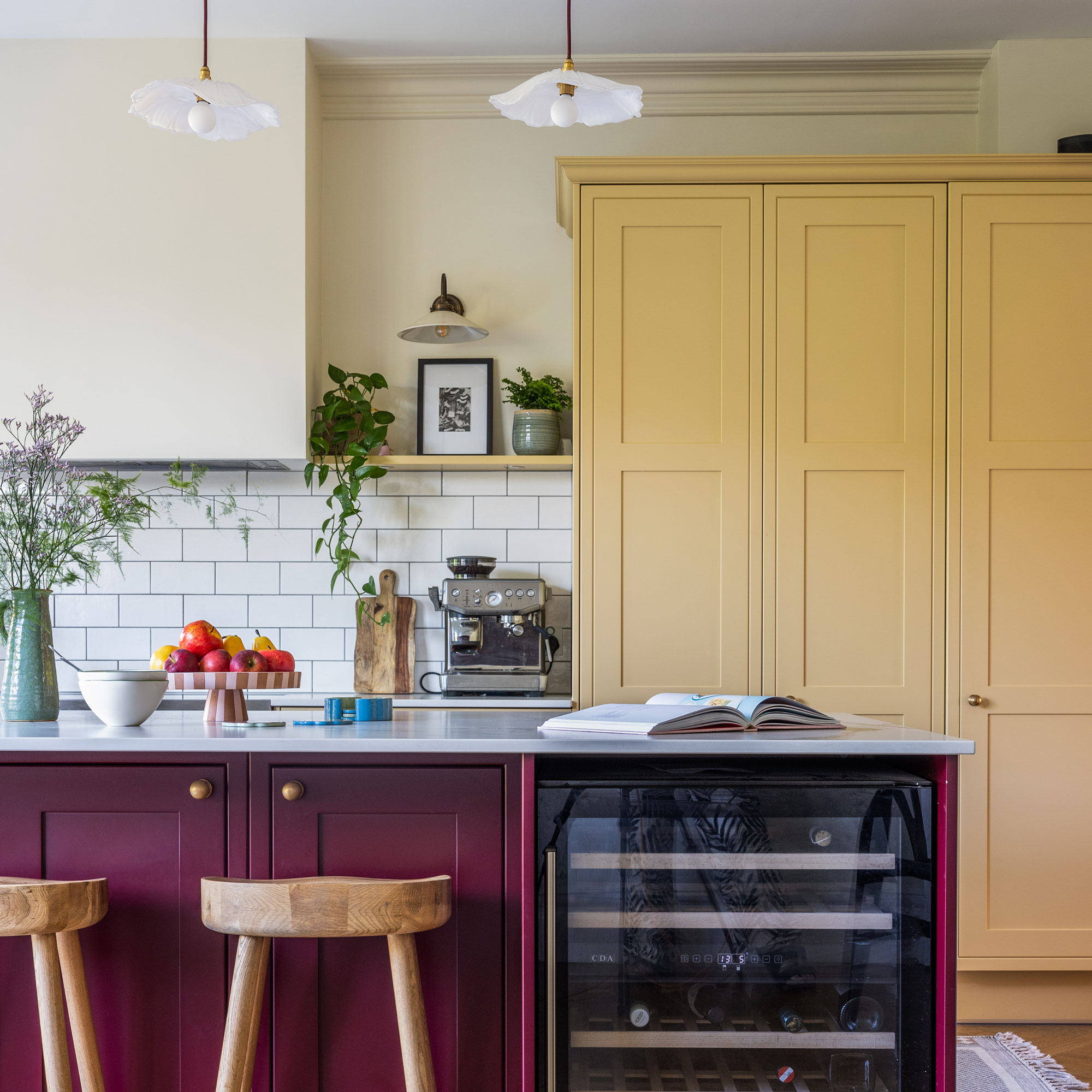 Yellow floor to ceiling kitchen cabinet, red kitchen island with wooden stools. A silver coffee machine sits on the white kitchen worktop.