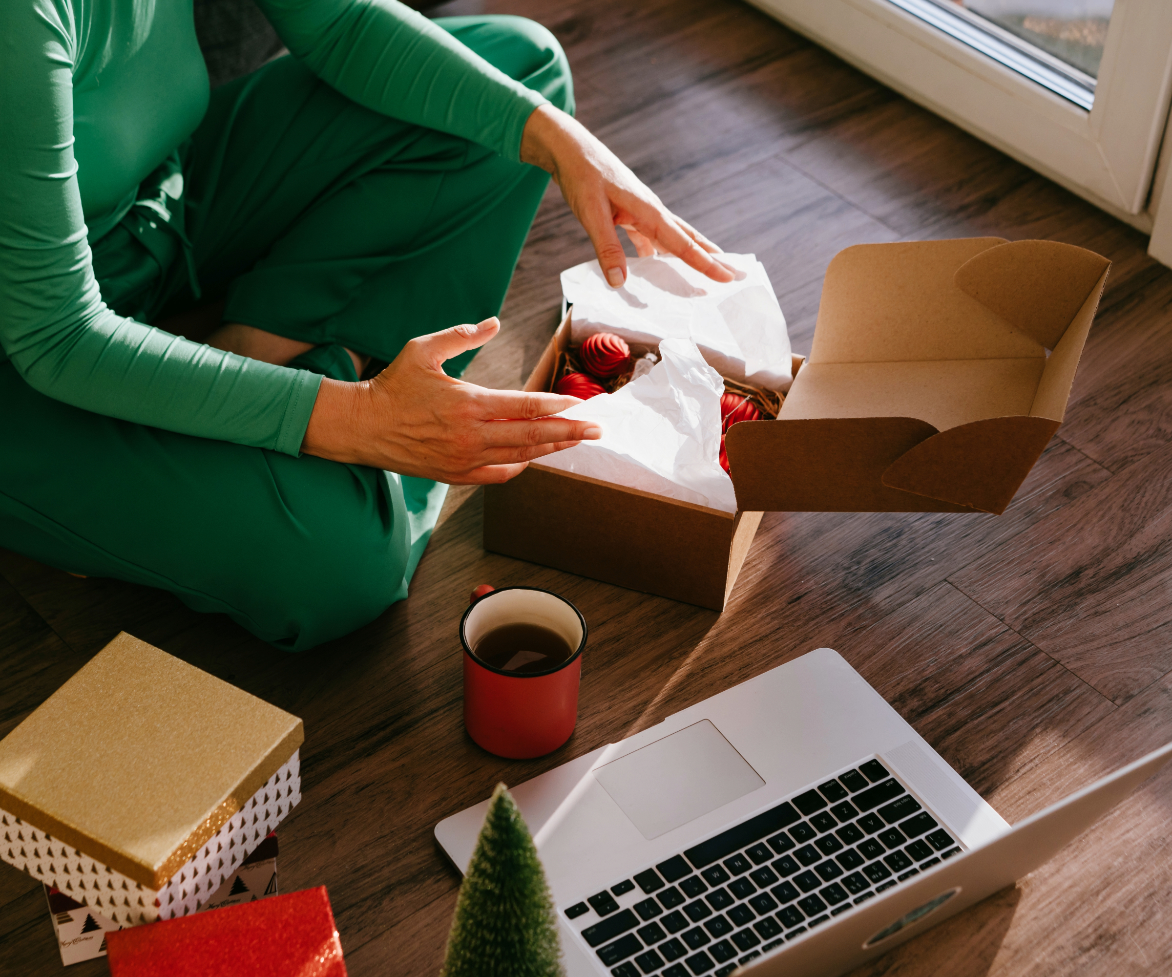 Person crossed legged with a laptop, mug of drink, and open gift box in front of them