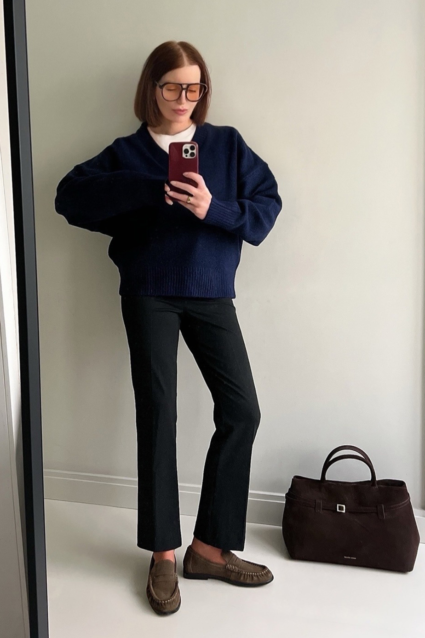 a woman wearing a navy sweater, black pants, and tan suede loafers with a brown suede bag