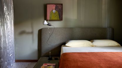 bedroom with green walls, green headboard, small artwork above bed, orange bedding, and silver curtain with light fitting hanging beside bed