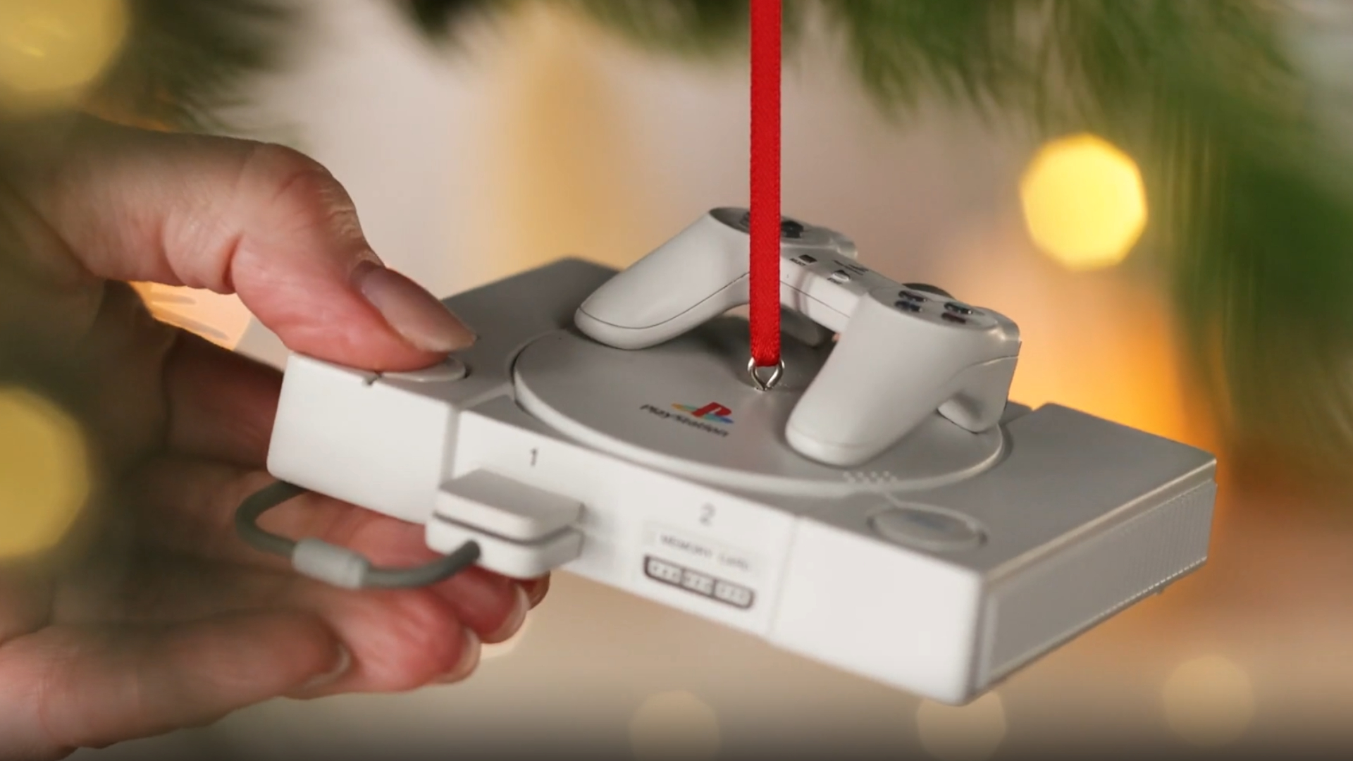 A hand presses the power button of a PlayStation ornament hanging from a Christmas tree