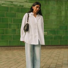 Woman wearing Madewell button-down white shirt, light wash jeans, and black shoulder bag stands in front of green wall.