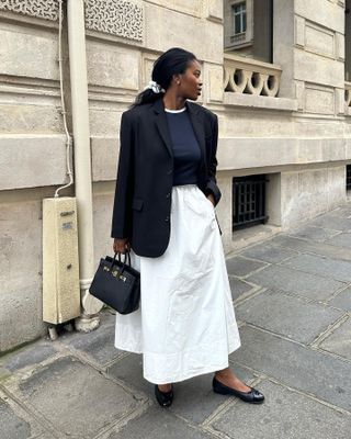 British fashion influencer Marilyn Nwawulor-Kazemaks poses on a London sidewalk wearing a low ponytail, a black blazer, layered basic tops, a full white skirt, an Hermes bag, and black ballet flats.