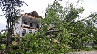 A tree fallen in front of a damaged house against a grey sky.