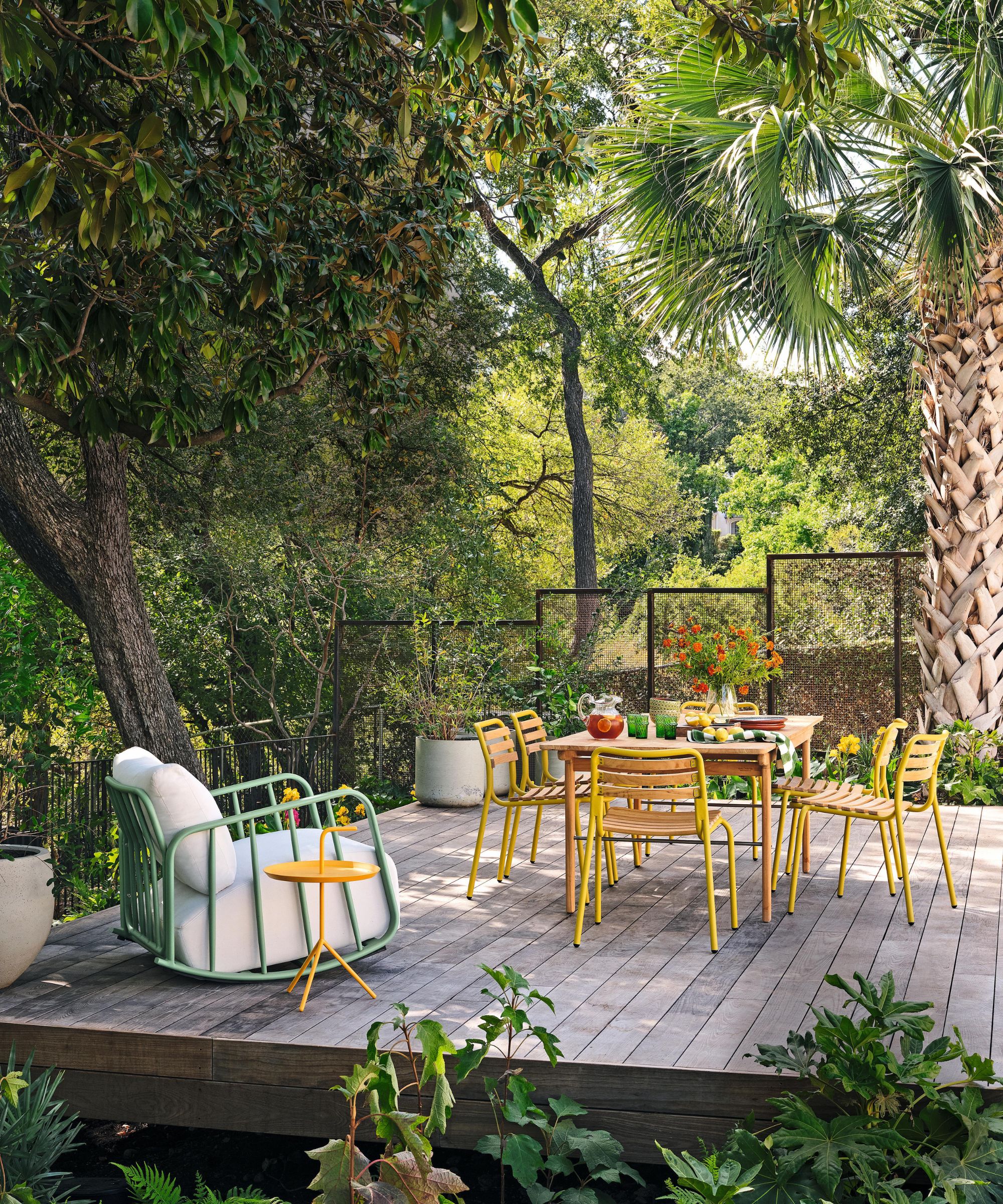 a leafy tropical inspired backyard with a deck area with yellow dining chairs and a green rocking chair