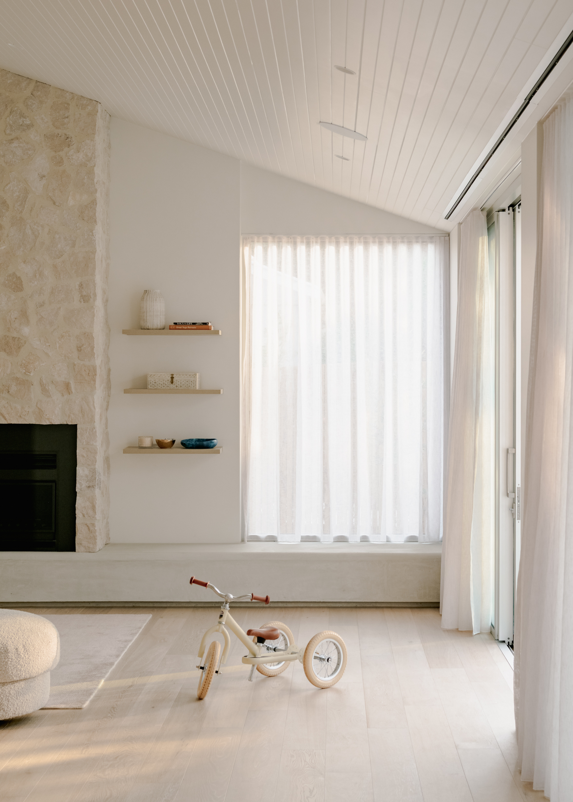 A minimalist living room with a white children's bike, a rug, a stone fireplace and three wall shelves