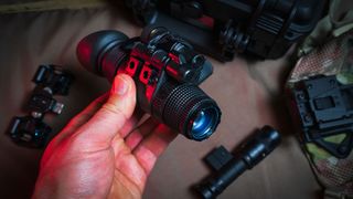 A hand holding a night vision monocular in front of optical gear.