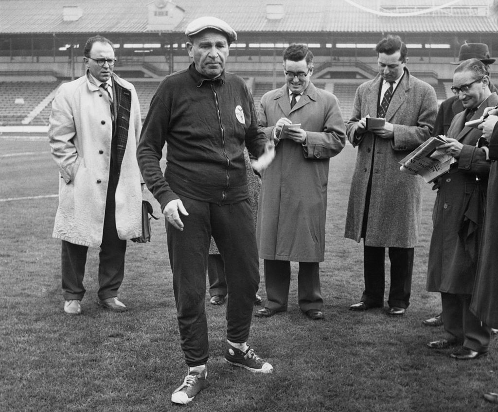 Benfica coach and manager Bela Guttmann (1900 - 1981) with a group of journalists at White City in London, 4th April 1962. He had just accused Tottenham Hotspur of watering the pitch at White Hart Lane, in preparation for their game against Benfica the next day. The match was the second leg semi-final of the European Cup.