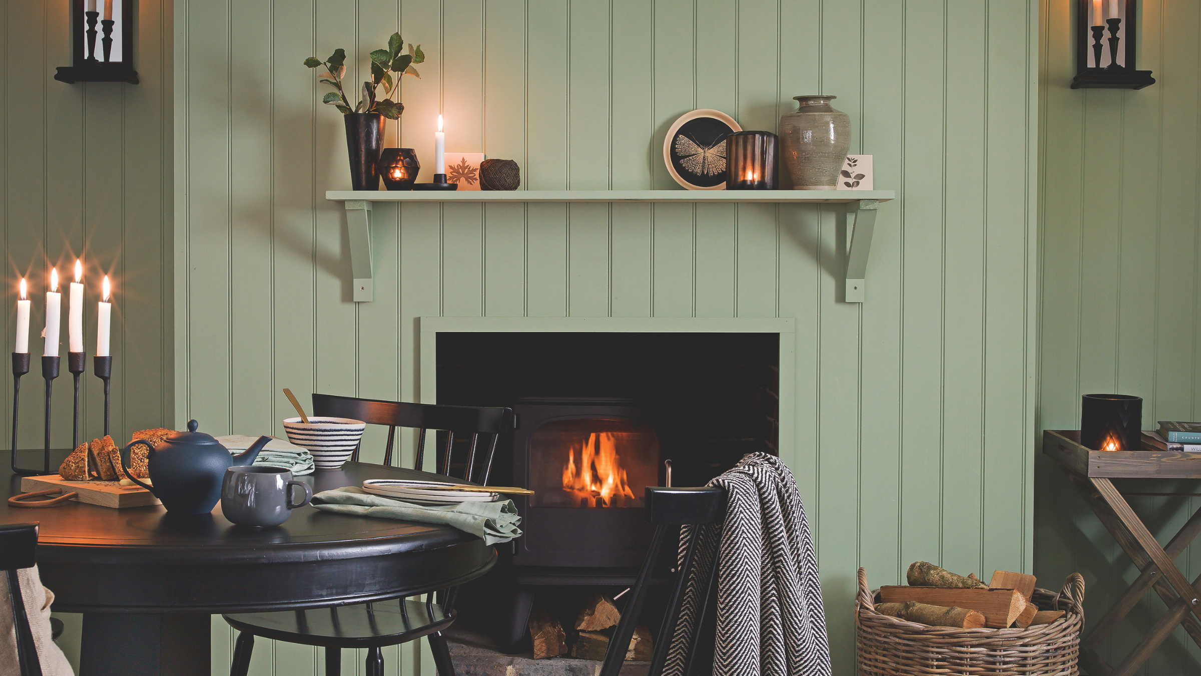 Cosy green panelled dining room with lit log burner, black dining table and chairs and candles.