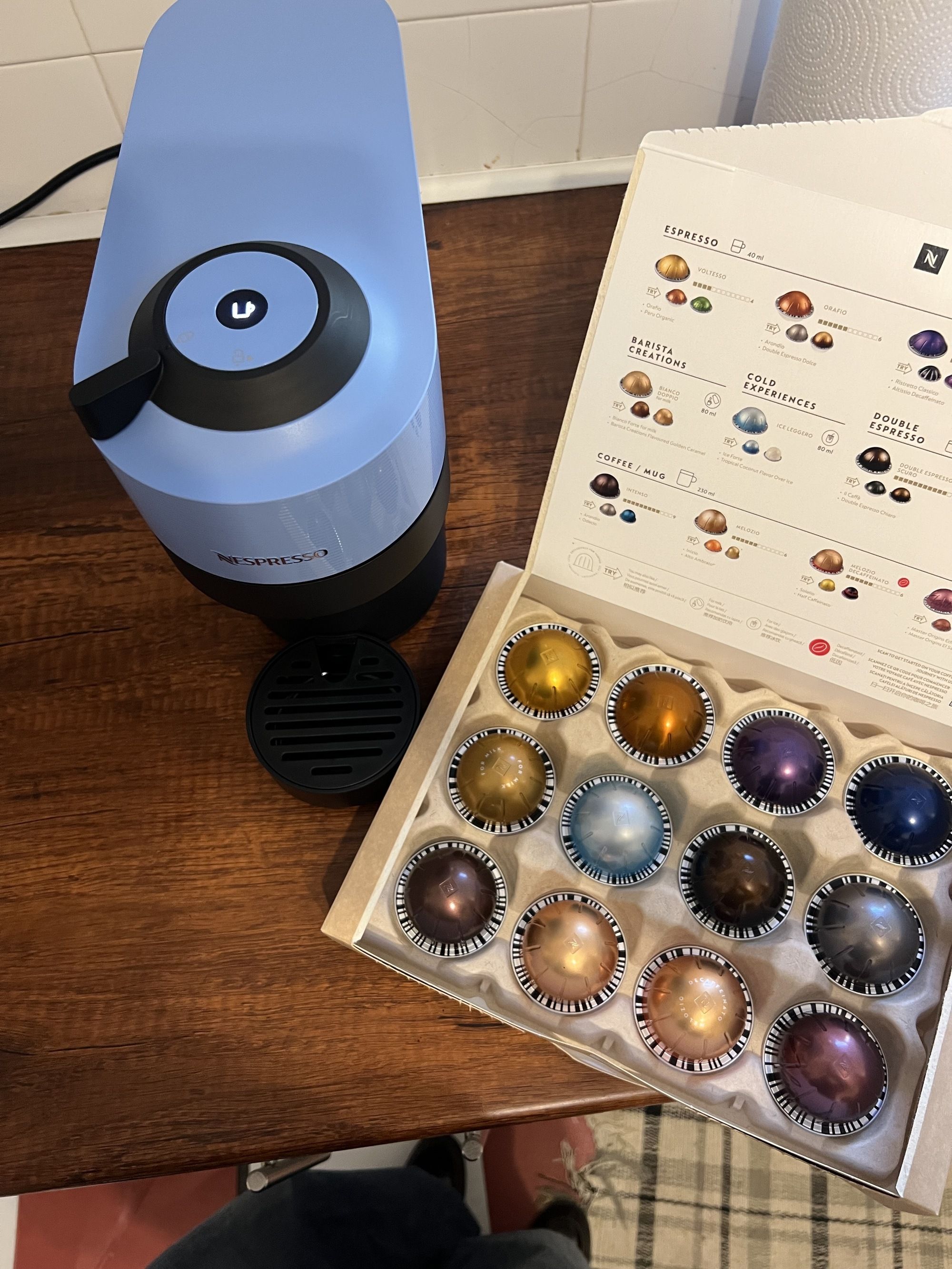 Image of a light blue and black Nespresso Vertuo Pop machine on a wooden kitchen countertop. There is a box of coffee pods in different colors and sizes next to the machine