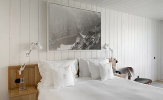 Bedroom with white linen with white cladded walls and ceiling. A black and white framed picture of the area is hanging on the wall.
