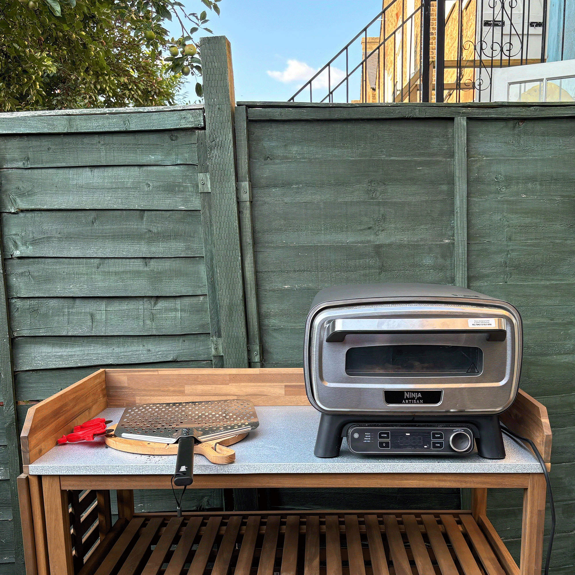 Ninja artisan pizza oven on top of a potting bench with a pizza peel and pizza board next to it