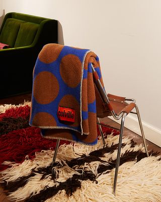 A room with a green sofa and a red, cream and brown shag rug with a brown chair on top. Placed over the chair is a folded blue Colville woolen blanket with brown spots and red tag